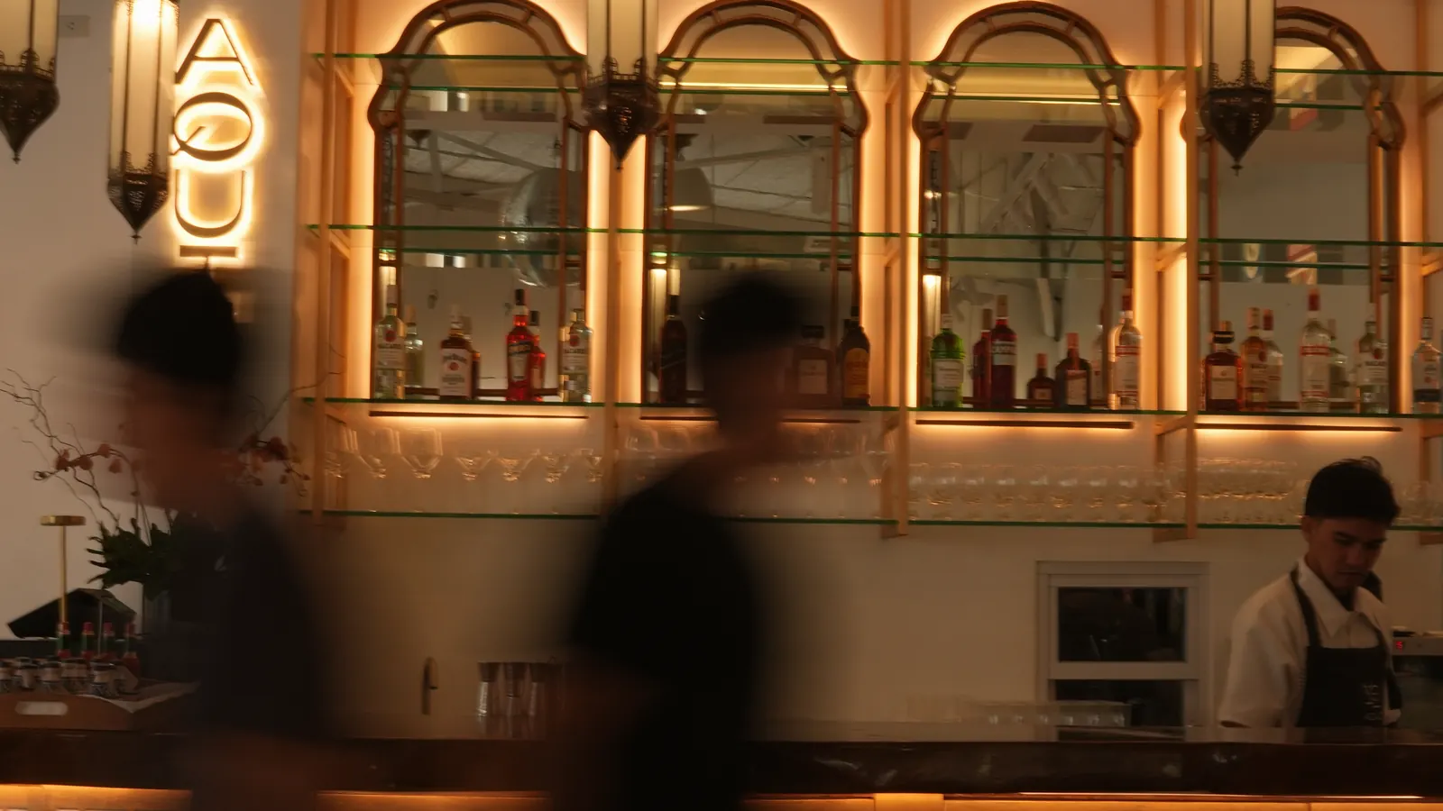 The Aqui bar at night: the lit AQUI sign, arched mirrors in gold frames and lanterns, two guests passing in a blur