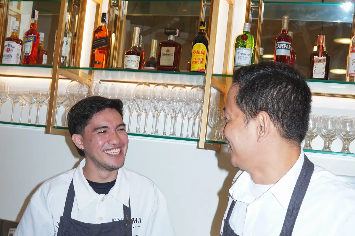 Two of the Aqui bar team in their AQUI aprons, laughing at the bar