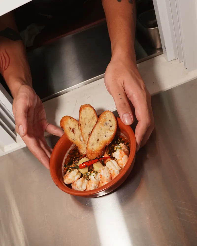 Two hands hold a dish of gambas al ajillo out through Aqui’s kitchen pass