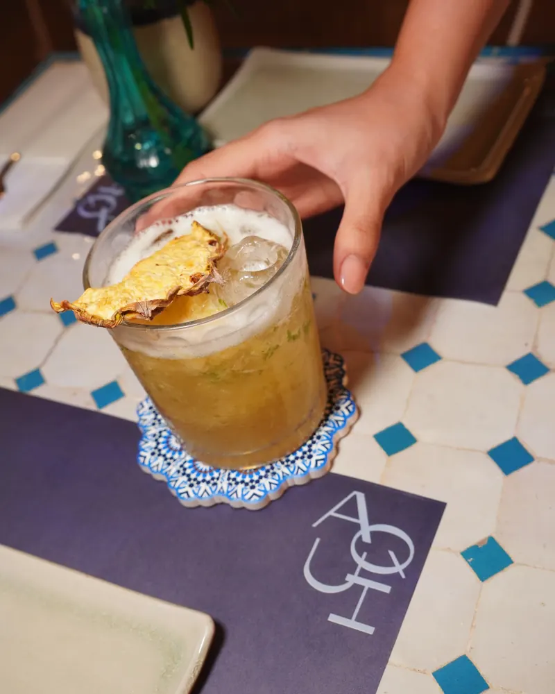 A Fuego de Piña with a dried pineapple slice, set down on its tile coaster on the AQUI placemat