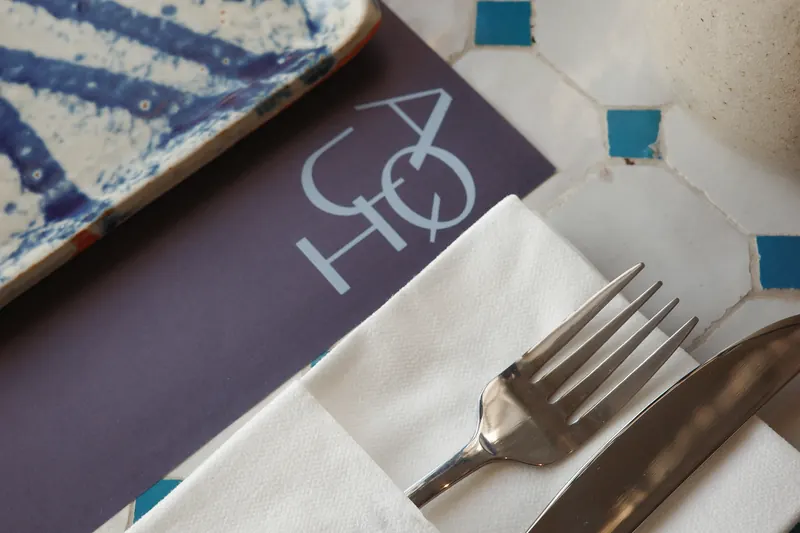 A place set at Aqui: the AQUI placemat, a folded napkin, fork and knife on the tiled table