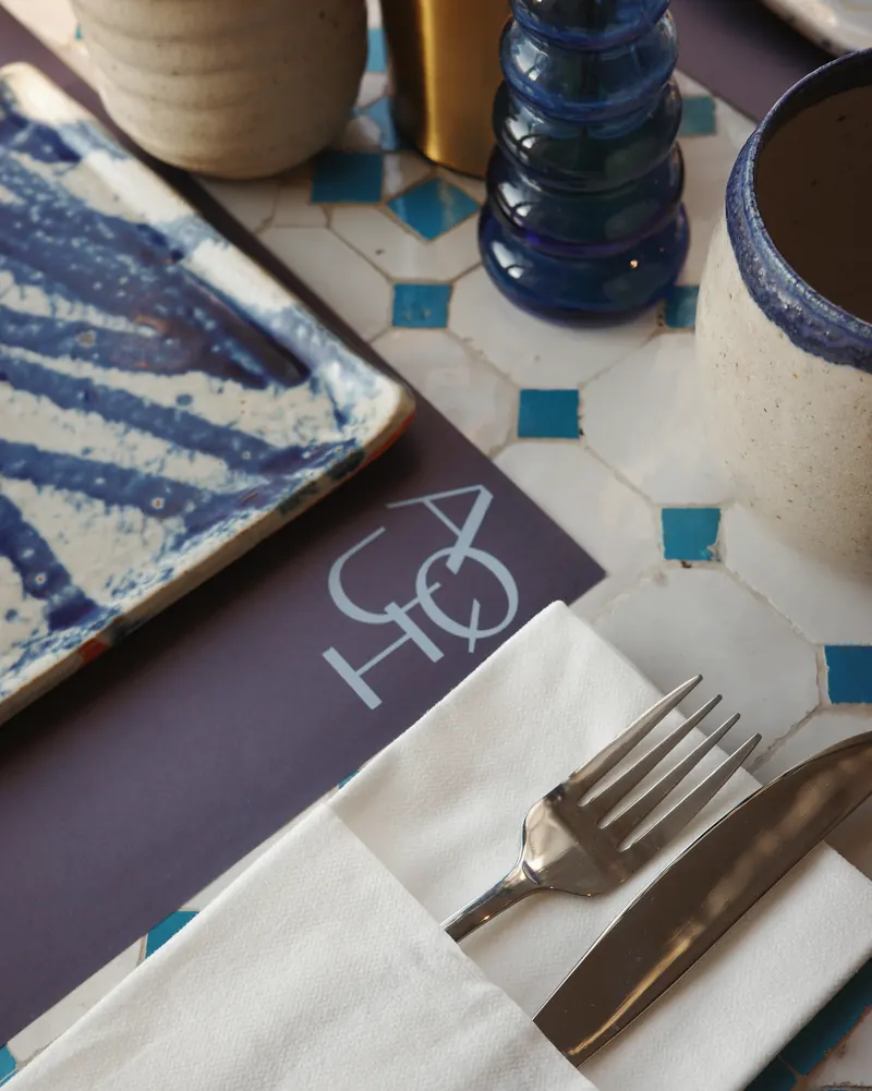 A place set on the tiled table: the AQUI placemat, a folded napkin, fork and knife, a blue-glazed plate