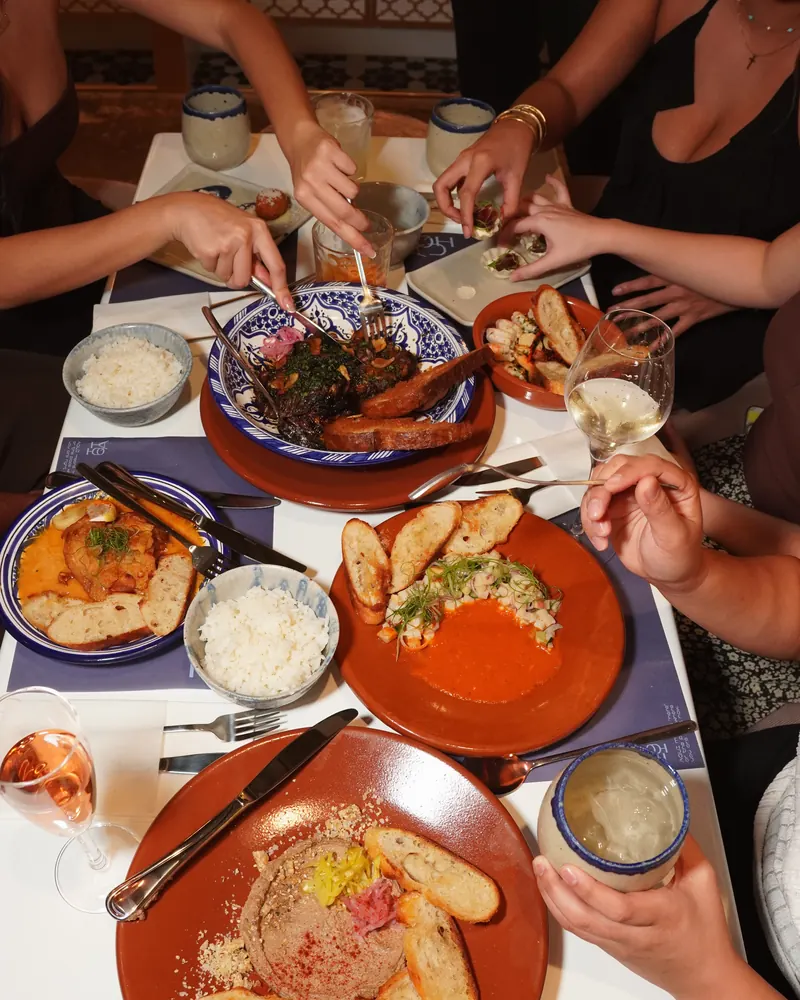 Hands reaching across a full table of small plates, rice and toasted bread
