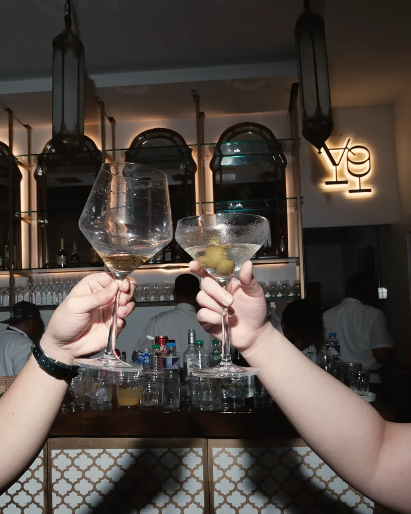 Two martinis raised in a toast at the Aqui bar, under its arched mirrors and the glowing cocktail-glass sign
